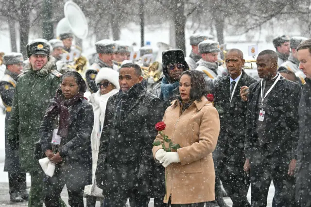 Легко одетая (без шапок, некоторые в пиджаках) мадагаскарская делегация в окружении российских военных, в том числе военного оркестра с трубами. Сверху падает снег, у президента Мадагаскара недовольно и немного растерянное лицо. Он одет в легкое пальто.