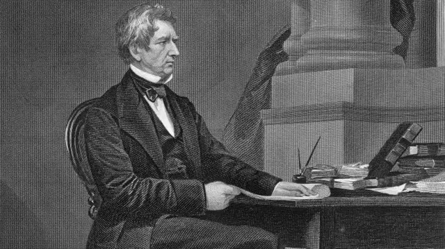 Un dibujo del secretario de Estado estadounidense, William Seward, quien negoció la compra de Alaska. Es en blanco y negro y muestra a un hombre sentado en una silla cerca de un escritorio. El hombre viste un elegante traje oscuro y apoya una mano en el escritorio. En el escritorio hay libros y papeles.