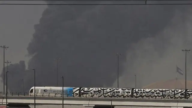 एक मार्च, 2026 को दुबई में कथित ईरानी हमले के बाद जेबेल अली बंदरगाह से उठते धुएं के सामने से गुज़रती एक मेट्रो ट्रेन