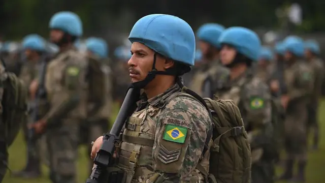 Um militar brasileiro usando uniforme camuflado, capacete azul e mochila aparece em primeiro plano segurando uma arma, enquanto outros militares com capacetes azuis permanecem perfilados ao fundo, desfocados.