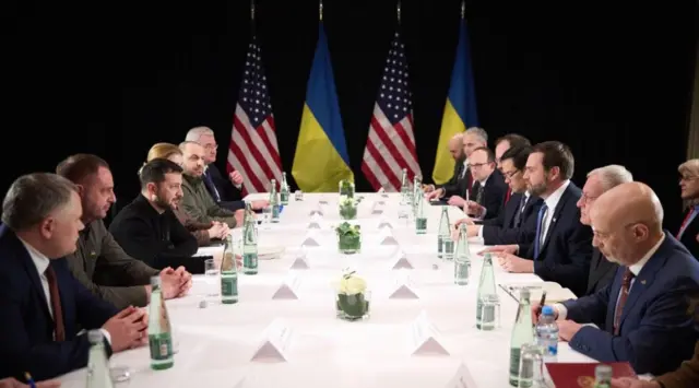 زيلينسكي وفانس على طاولة اجتماعات مع عدد من المسؤولين من الجانبين، بجانب طرف الطاولة 4 أعلام اثنان للولايات المتحدة واثنان لأوكرانيا. على الطاولة أسماء الحضور وزجاجات مياه