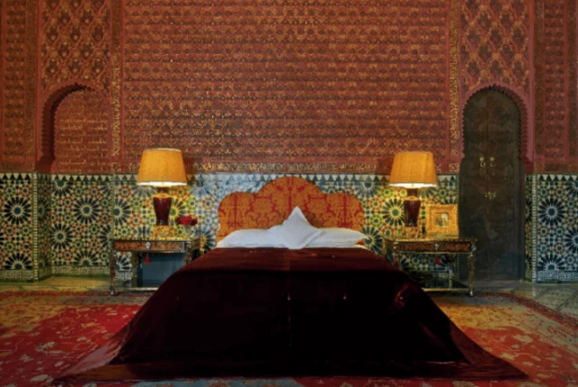 Zdjęcie wnętrza sypialni z łóżkiem nakrytym ciemnoczerwoną narzutą. Pokój jest bogato zdobiony kafelkami i czerwonymi rzeźbionymi ścianami.