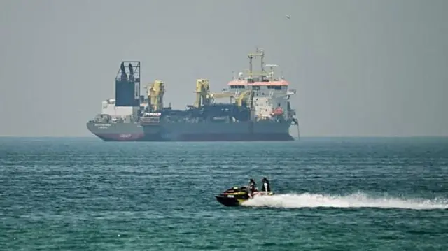 সাগরে ভাসমান একটি বাণিজ্যিক জাহাজ, কিছুটা দূরে একটি ওয়াটার বাইক দেখা যাচ্ছে; ২০শে এপ্রিল ২০২৬ এর ছবি