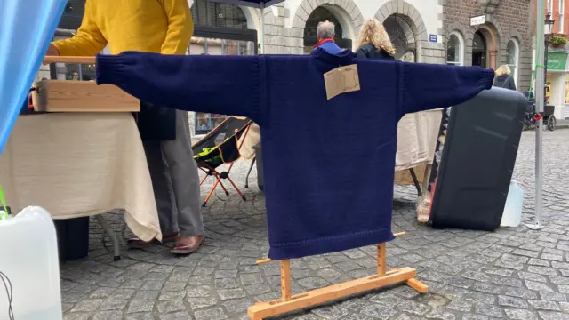 Un jersey azul de punto de Guernsey cuelga de un soporte con las mangas extendidas. El soporte está sobre adoquines grises. Detrás, un hombre con pantalones grises y un jersey amarillo de Guernsey está de pie. Hay una mesa y una pérgola. Al fondo se ven tiendas.