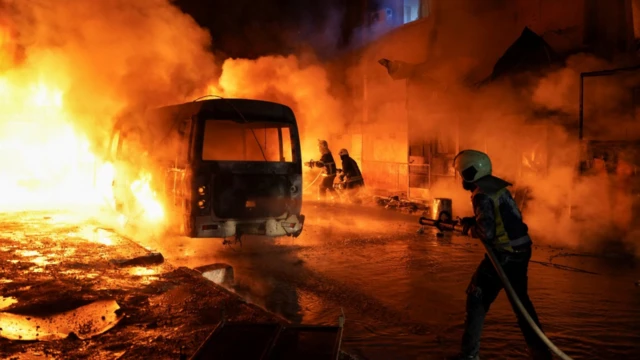 Üç itfaiye görevlisi alev alev yanan bir otobüsü su sıkarak söndürmeye çalışıyor. Gece karanlığında ortam alevlerin ışığıyla aydınlanmış durumda.