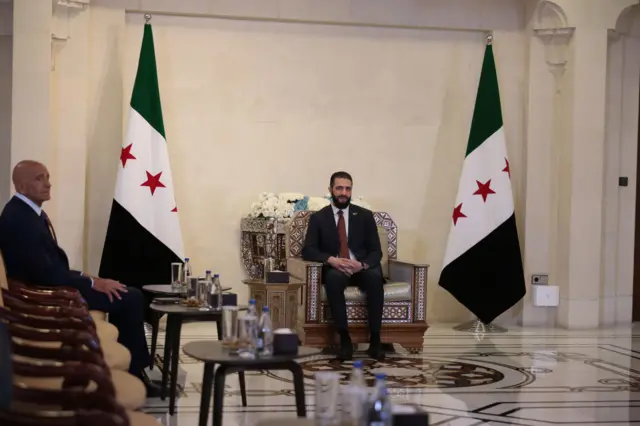 Barrack ve Eş-Şara'nın görüşmesinden bir fotoğraf. Eş-Şera Suriye bayraklarının ortasında oturuyor. Barrack ise sağında en yakın sandalyede oturuyor.