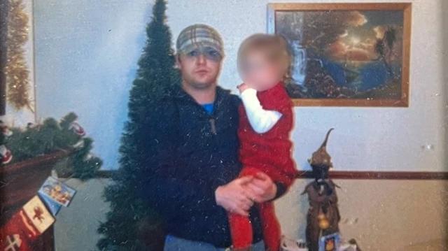 Un hombre con gorra a cuadros y sudadera azul marino con cremallera, sosteniendo a un niño vestido de rojo. De pie frente a un árbol de Navidad en una sala de estar.