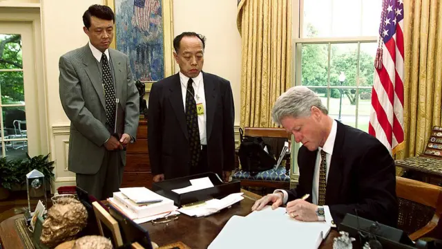 সমবেদনা বইয়ে লিখছেন প্রেসিডেন্ট ক্লিন্টন, পাশে আমেরিকায় চীনা রাষ্ট্রদূত ও একজন চীনা কর্মকর্তা - ১৩ই মে ১৯৯৯