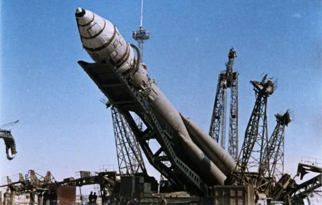 Büyük bir roketin fırlatma için vinçler tarafından kuleye dik olarak yerleştirilme anını gösteriyor. Roket fotoğrafta henüz eğik durumda.