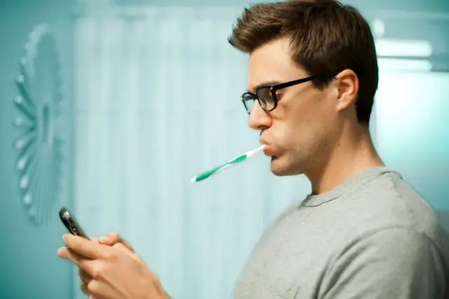 Un hombre de perfil en un baño, con anteojos y un cepillo de dientes en la boca, consulta su teléfono móvil