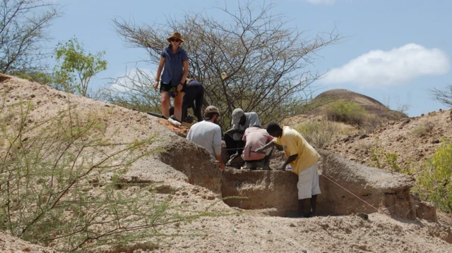 A group of people are working together at an archaeological dig site in a dry, rocky landscape with sparse vegetation. Some are kneeling and carefully examining or excavating a partially exposed layer of earth, while one stands nearby watching. Their focused activity suggests they are searching for or studying artifacts or fossils in the ground.