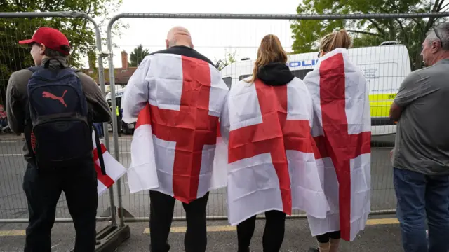 金属フェンスの後ろに5人の人物が立っている。うち3人は肩にイングランドの旗をかけている。彼らの前にはベル・ホテルがあり、警察車両によって封鎖されている