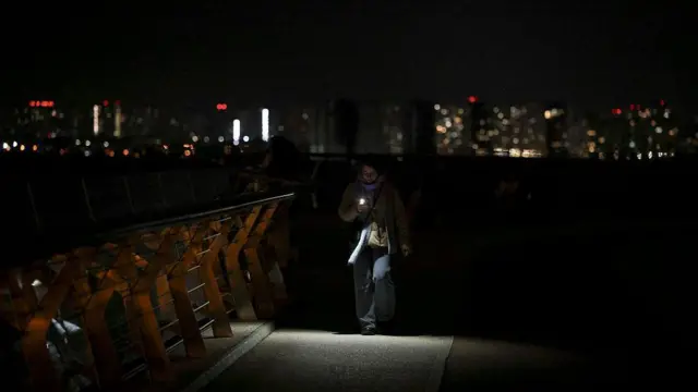 Київ під час блекаутів. Дівчина йде в темному місті, підсвічуючи собі телефоном, і далі далеко позаду видно вогні міста