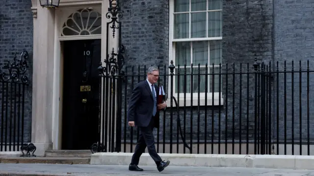 施紀賢爵士步出倫敦唐寧街10號首相府後往街口走去，左手抱著兩個文件夾（26/11/2025）