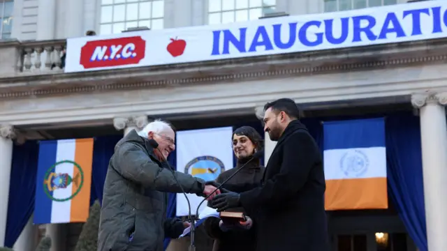 「ニューヨーク市 りんごマーク 就任式」の横断幕や、紺・白・オレンジの州旗が掲げられた、灰色の市役所の建物の前で、黒いコートに手袋のマムダニ氏とグレーのブルゾンを着た白髪のサンダース氏が笑顔で握手している。間に、宣誓用のコーランを手にしたマムダニ夫人のラマ・ドゥワジ氏が黒いコート姿で笑顔を浮かべている