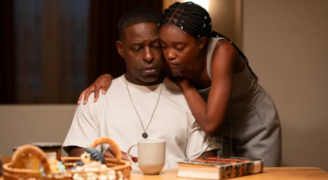 Un hombre, sentado en una mesa, con una taza y un libro delante, con una joven que lo abraza por detrás. 