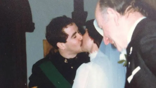 Há quatro pessoas nesta fotografia. No centro, um casal de noivos se beija no dia do casamento. O noivo veste um uniforme militar preto. A noiva, de costas para a câmera, usa um vestido branco de gola alta e um véu branco preso a um adorno de cabeça. Ambos têm cabelos escuros. À esquerda do noivo está o cônego, que celebrou o casamento. À direita da noiva, um convidado do casamento veste um terno preto.