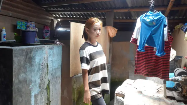 Esti Agustina mengenakan baju garis-garis hitam putih dan celana hitam, berdiri menyamping di dalam tempat yang tampak seperti kamar mandi luar ruangan.