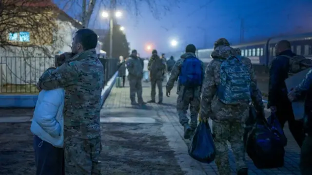 Пара обнимается на платформе железнодорожного вокзала, мимо проходят военные