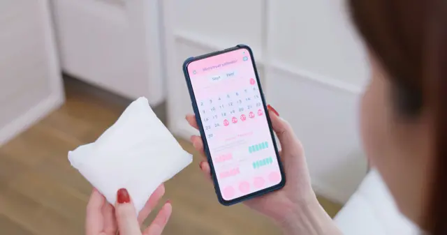 Une femme tient un téléphone portable avec une application de suivi des règles à l'écran, tandis que dans l'autre main elle tient une serviette hygiénique.