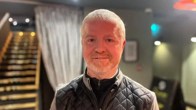 Keith aparece en un bar. El fondo está borroso, pero se ven unas escaleras. Keith tiene el pelo corto y blanco, un chaleco negro y un jersey negro con cremallera tres cuartos.