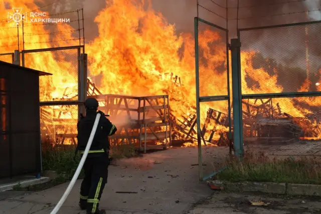 Пожежа на відкритій території: рятувальник зі шлангом наближається до великих вогняних язиків, які охопили складені дерев’яні конструкції за металевою огорожею. Полум’я високе й інтенсивне, навколо видно задимлення.