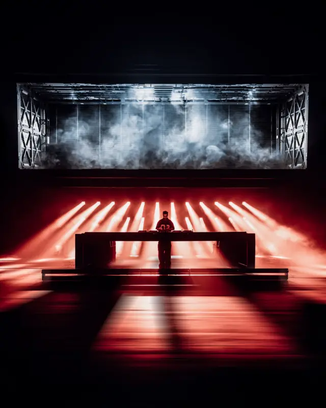Alok sozinho no centro de um palco, atrás de uma ampla mesa de som, iluminado por feixes de luz vermelha que se projetam em direção ao público. Ao fundo, uma grande estrutura metálica envolta por fumaça e iluminação branca cria uma atmosfera futurista e industrial.