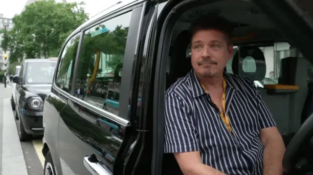 Paul Brennan, presidente da Associação de Motoristas de Táxi Licenciados, sentado ao volante de um táxi preto. Ele tem uma expressão perplexa e veste uma camisa azul-marinho com listras verticais brancas. Atrás dele, há outro táxi preto estacionado na calçada.