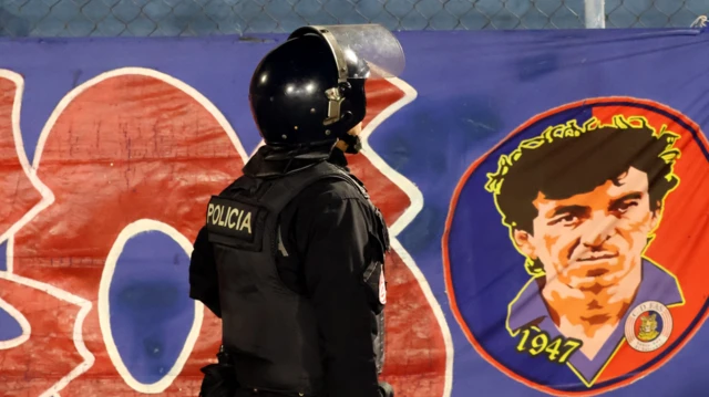 Policial em primeiro plano, de costas, frente a um cartaz com uma figura do futebol salvadorenho, o Mágico González, e o ano de 1947, no Estádio Óscar Alberto Quiteño, onde o time local do FAS enfrentou o Municipal Limeño, em Santa Ana (El Salvador), no dia 11 de outubro de 2025