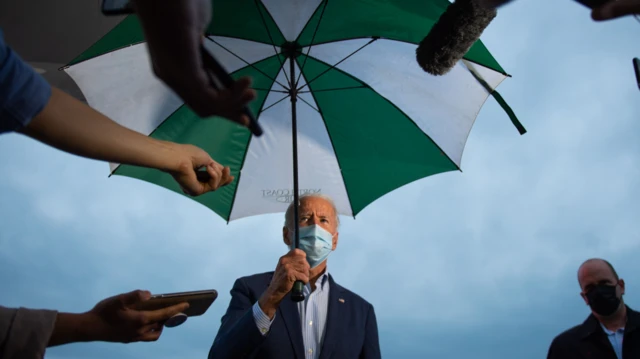 Joe Biden under an umbrella with a mask on, as press ask questions 