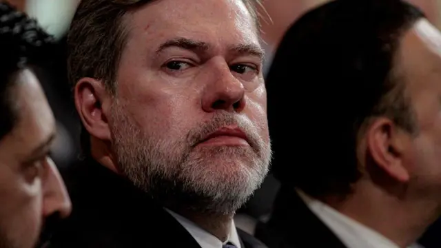 O ministro José Dias Toffoli, do STF, um homem de meia-idade, com barba grisalha e expressão séria, olha de lado. Ele veste terno escuro e camisa social, e está sentado entre outras pessoas, que aparecem desfocadas ao redor. A iluminação destaca seu rosto.