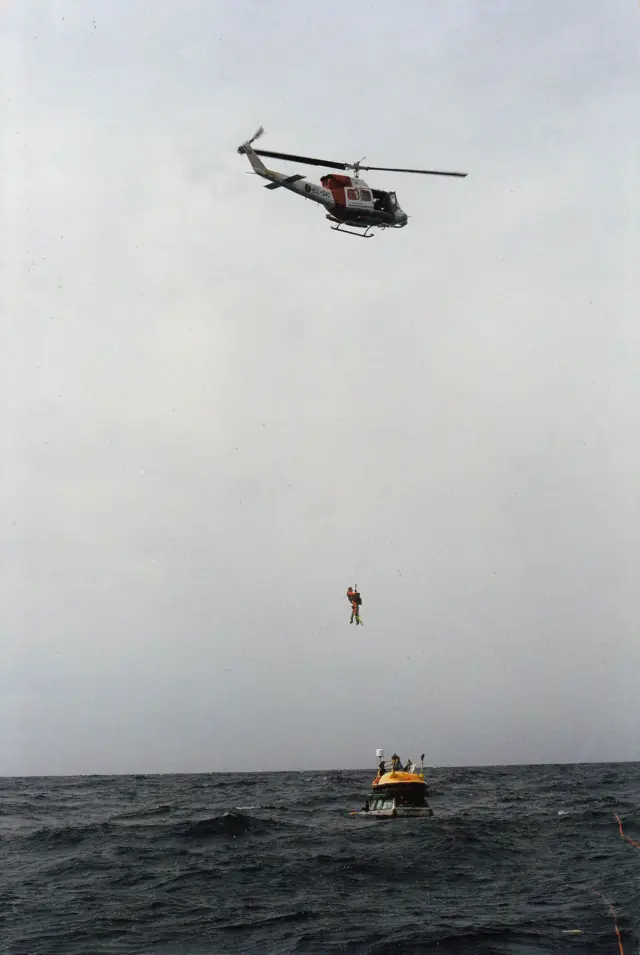 Denizdeki arabaların üzerinde bir helikopter havada asılı duruyor ve iki adamı güvenli bir yere taşıyor.