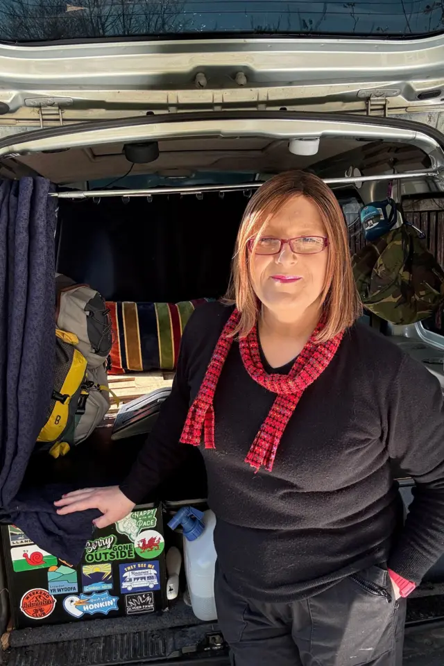 Жінка з волоссям до плечей стоїть біля відкритого заднього відсіку фургона, переобладнаного для практичного використання на природі. Вона в чорній кофті та червоному шарфі з візерунком, спирається рукою на край фургона, інша рука в кишені. 