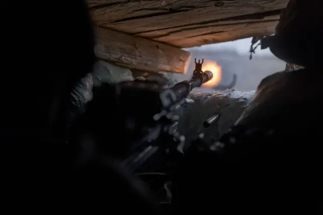 Знімок зсередини бліндажа з дерев’яним перекриттям угорі. У центрі кадру видно ствол зброї, спрямований назовні. На самому кінці ствола спалахує яскравий вогняний спалах – момент пострілу. Праворуч і ліворуч у тіні проглядаються силуети двох людей, які займають позиції всередині укриття. 