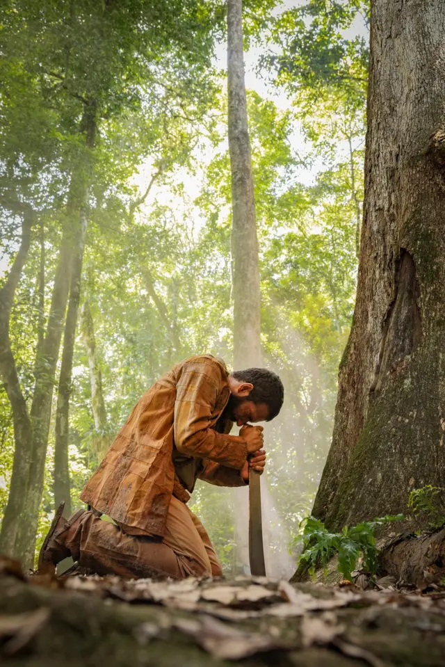 A imagem mostra um homem ajoelhado na floresta, de cabeça baixa, apoiando as mãos sobre o cabo de um facão fincado no chão. Vestindo roupas sujas de terra, ele aparece em um momento de aparente exaustão ou reflexão, enquanto feixes de luz atravessam a copa das árvores e iluminam a cena, reforçando o ambiente denso da mata.