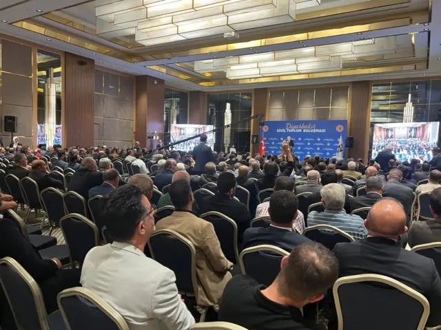 Kurtulmuş büyük bir salonda kürsüde. Odakta sandalyede oturan katılımcılar var.