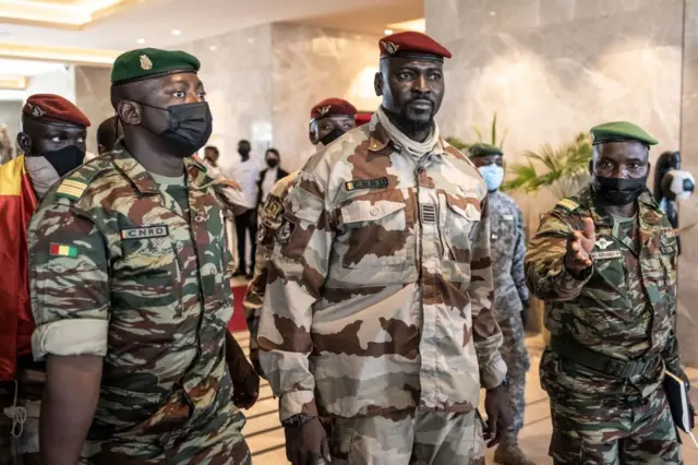 Mamadi Doumbouya, en uniforme militaire avec un béret rouge sur la tête, il est entouré de chaque côté par deux hommes, aussi en tenue militaire portant des bérets verts.