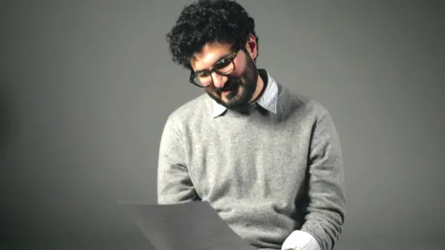 一個有著濃密捲曲的黑髮、鬍鬚和絡腮鬍的男人正在看著一張紙。他戴著眼鏡，穿著淺灰色套頭衫，內搭淺色襯衫，下身是淺棕色長褲。