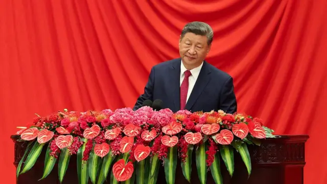Xi Jinping, vestido con un traje oscuro, se encuentra frente a una cortina roja y detrás de un atril de madera cubierto de flores rojas y rosas brillantes y hojas verdes.