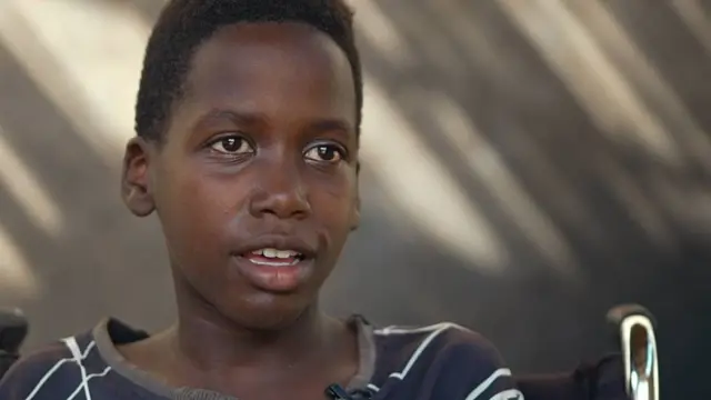 Zaher mirando y hablando a la cámara. El niño está sentado en una silla de ruedas y viste una camiseta negra gastada con rayas blancas.