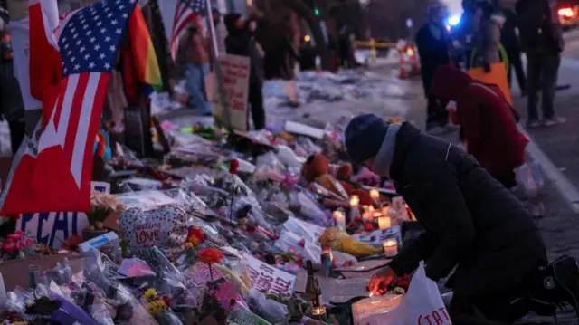 たくさんの花束やメッセージカード、アメリカなど複数の国の国旗が並ぶ。日が落ち始めた薄暗がりに、ろうそくの光が目立つ。複数の人が路上に膝をつき、ろうそくに火をともしたり、うつむいたりしている