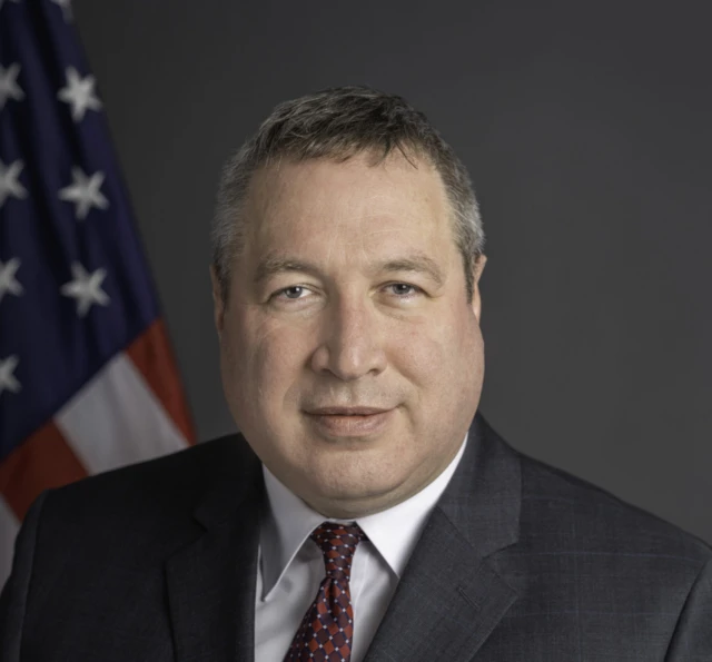 David Gamble usando terno em frente uma bandeira dos Estados Unidos