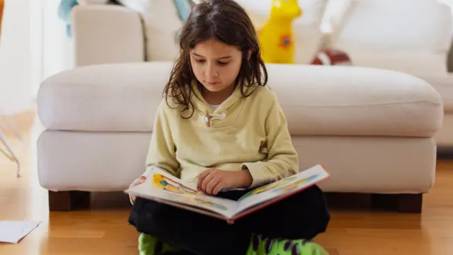 Sześciolatka czyta książkę, siedząc na drewnianej podłodze w salonie.