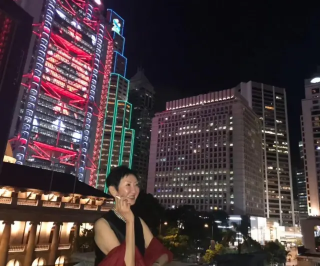 身穿晚装的施南生摄于香港会，日期不详，背景是中环夜景，从左至右分别是中国香港特区终审法院、汇丰银行香港总行、渣打银行香港总行和香港文华东方酒店