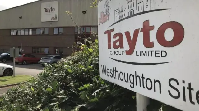 Un cartel afuera de una fábrica dice "Tayto Group Ltd Westhoughton site", con varios autos estacionados frente al edificio.