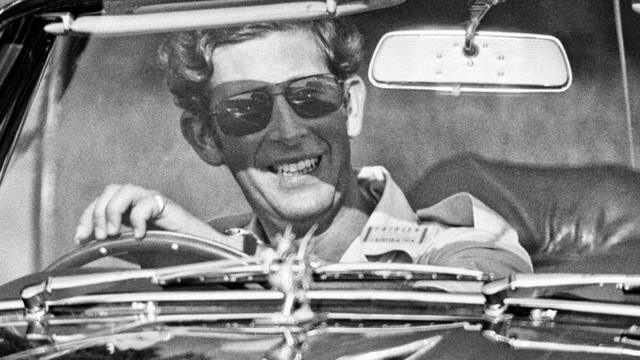 Rei Charles 3º usando óculos escuros e sorrindo dentro do esportivo Aston Martin em Windsor, Inglaterra, em 1975