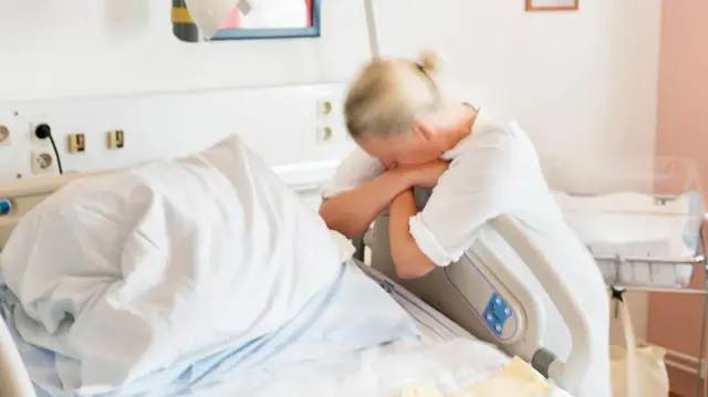 Obinrin kan fi ara ti bẹẹdi ileewosan, o wọ aṣọ funfun. O gbori le ọwọ rẹ mejeeji. 