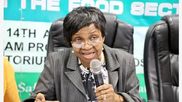 Aworan Ọga agba ajọ NAFDAC, Ọjọgbọn Mojisola Adeyeye lasiko to n ṣalaye ọrọ. Mama naa wọ aṣọ oyinbo alawọ eeru, o wọ funfun to ni dudu diẹ sinu o si fi ilẹkẹ sọrun, o lo yẹri eti funfun. Ọjọgbọn Adeyeye mu ẹrọ gbohun-gbohun lọwọ, oruka wa ni ọmọ ika rẹ kẹrin