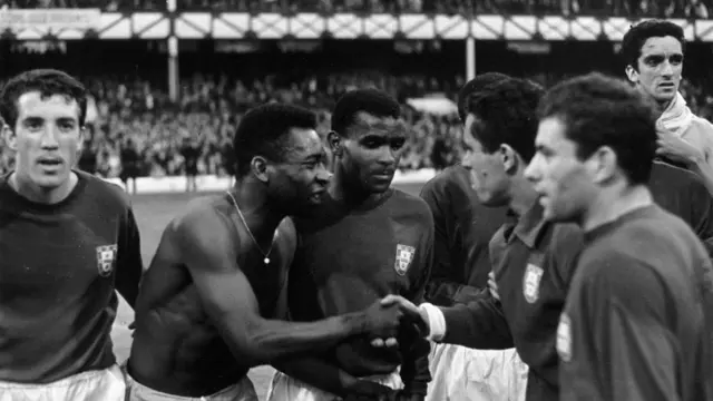 A imagem em preto e branco mostra jogadores das seleções de Brasil e Portugal reunidos após uma partida. Em primeiro plano, um jogador brasileiro sem camisa aperta a mão de um atleta português, enquanto outros jogadores observam a cena, em um registro que transmite respeito e confraternização entre as equipes diante de um estádio lotado.