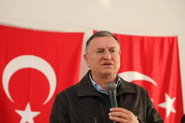 Lütfü Savaş iki Türkiye bayrağının önünde mikrofona konuşuyor. 